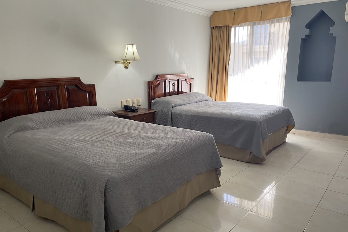 Habitación de hotel con dos camas, una mesita de noche, una lámpara y una ventana con cortinas.