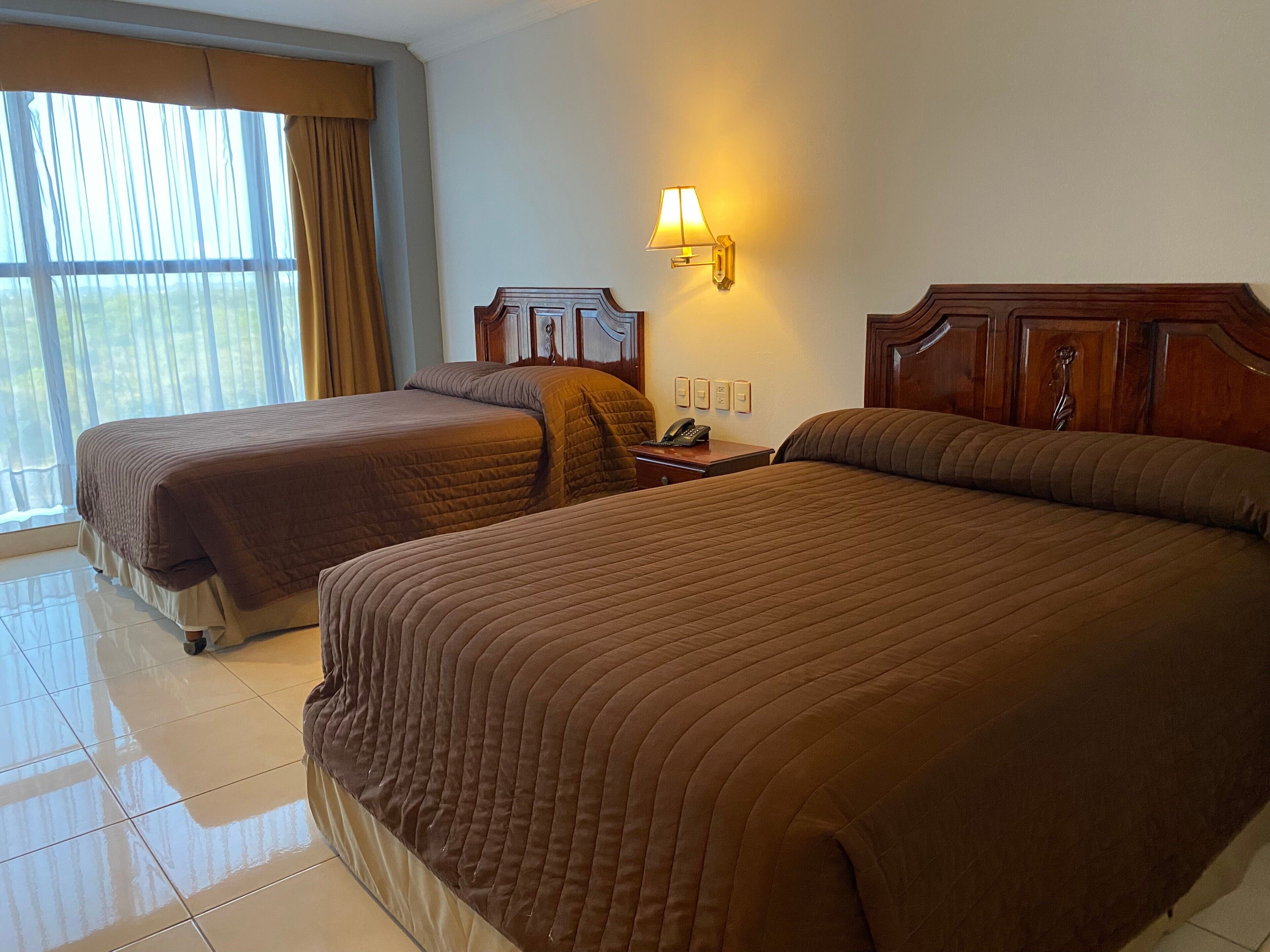 Una habitación de hotel con dos camas, una mesita de noche con lámpara y una ventana con cortinas.
