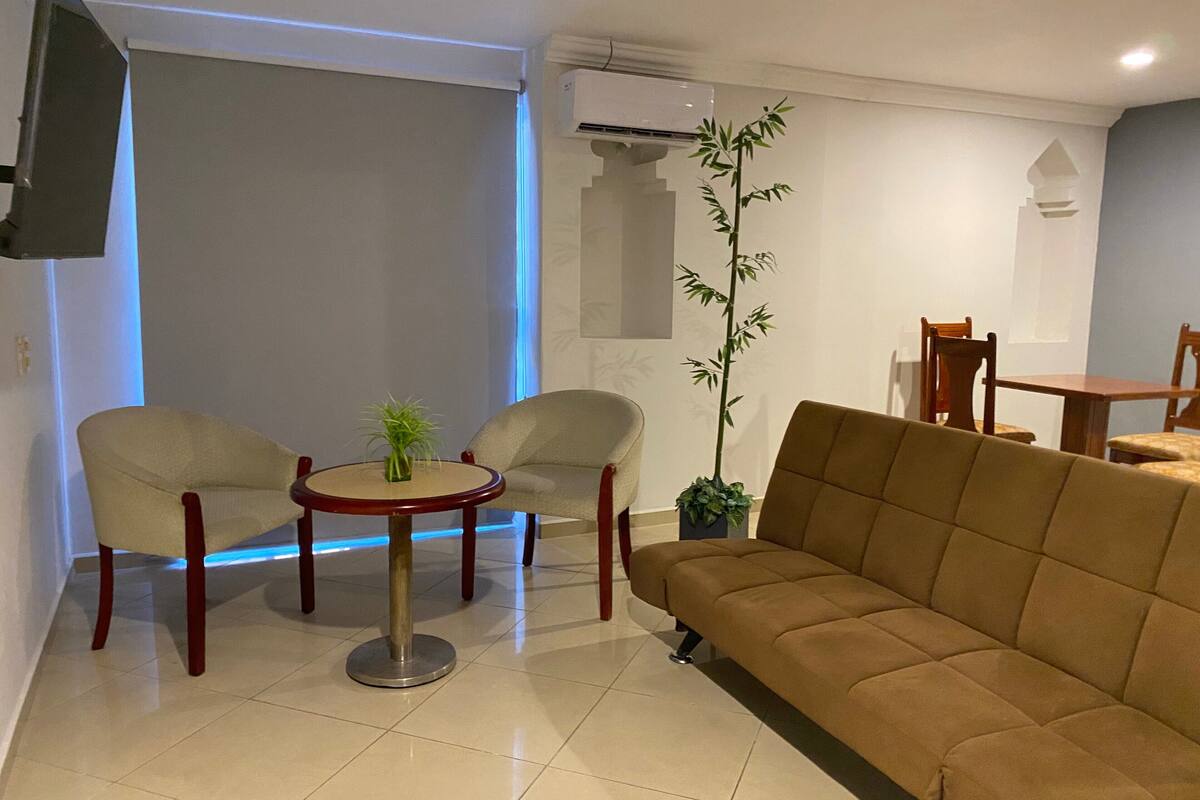 Una sala de estar con un sofá, dos sillones, una mesita redonda con una planta, y un comedor con mesa y sillas.