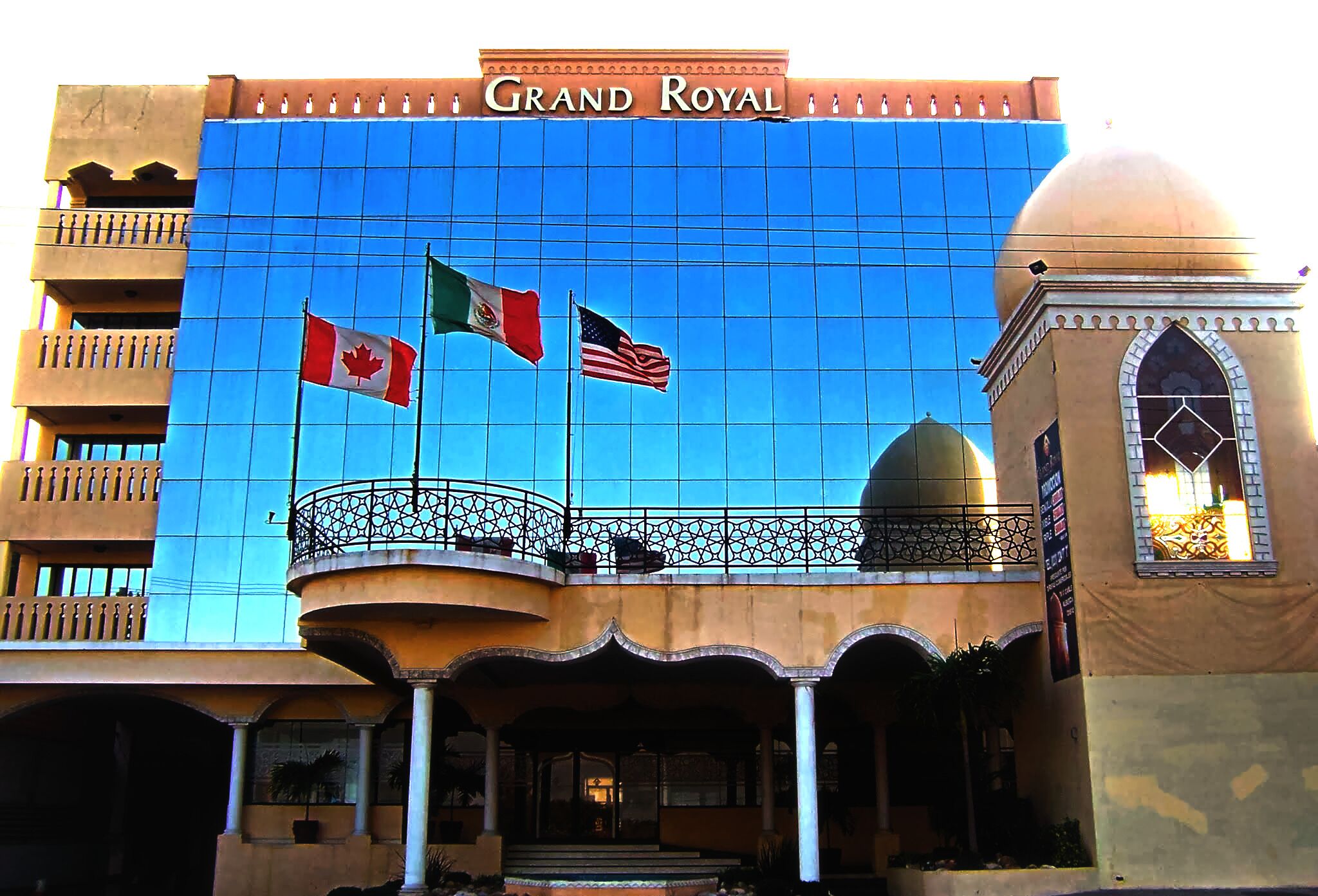 Un hotel con una amplia fachada de vidrio azul, una entrada destacada con balcón y banderas de Canadá y los Estados Unidos.