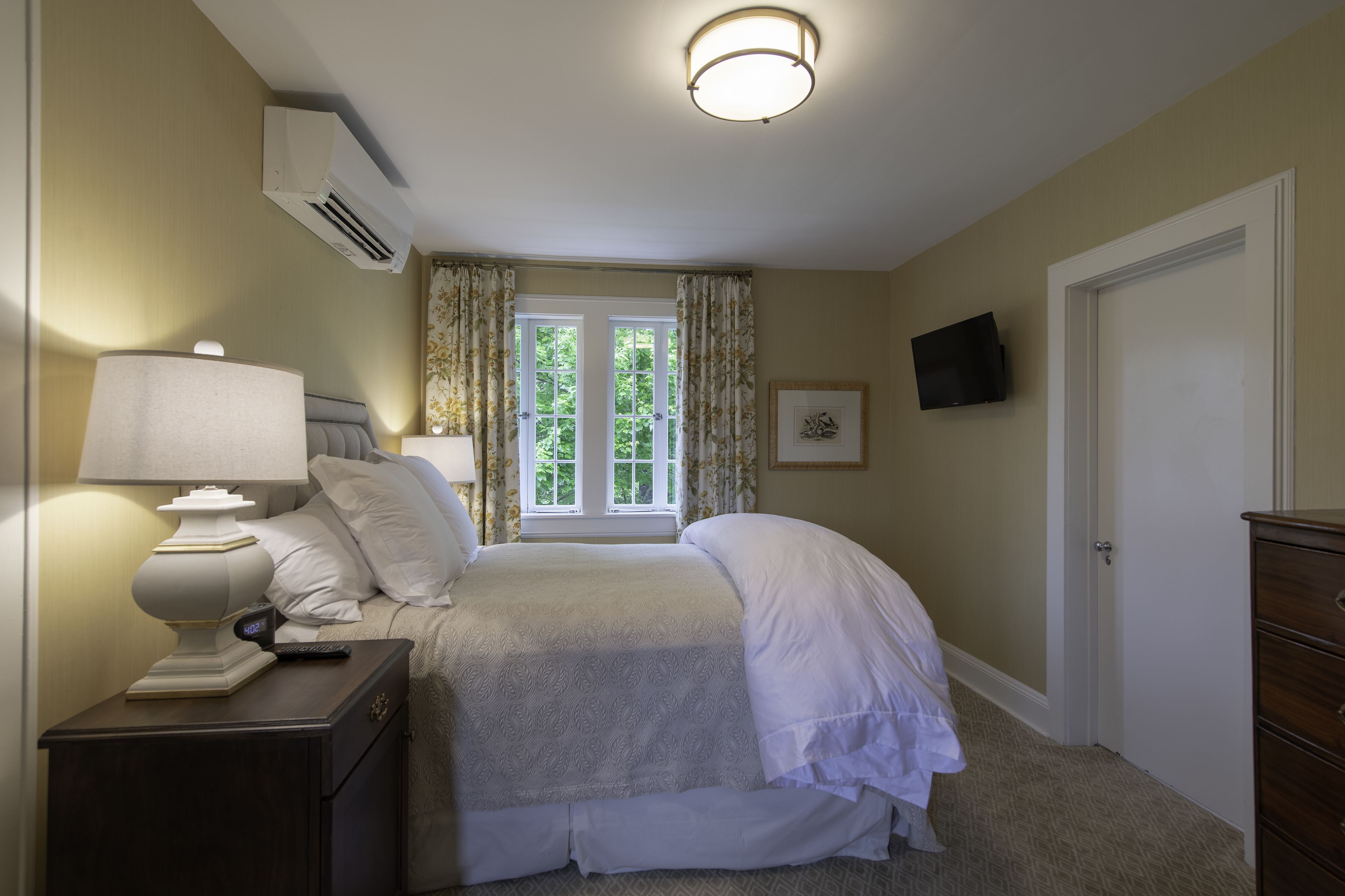 A bedroom with a bed, nightstand, lamp, air conditioning unit, window with curtains, and a television on the wall.