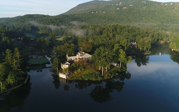 A large house surrounded by a lake with a forested hill in the background.