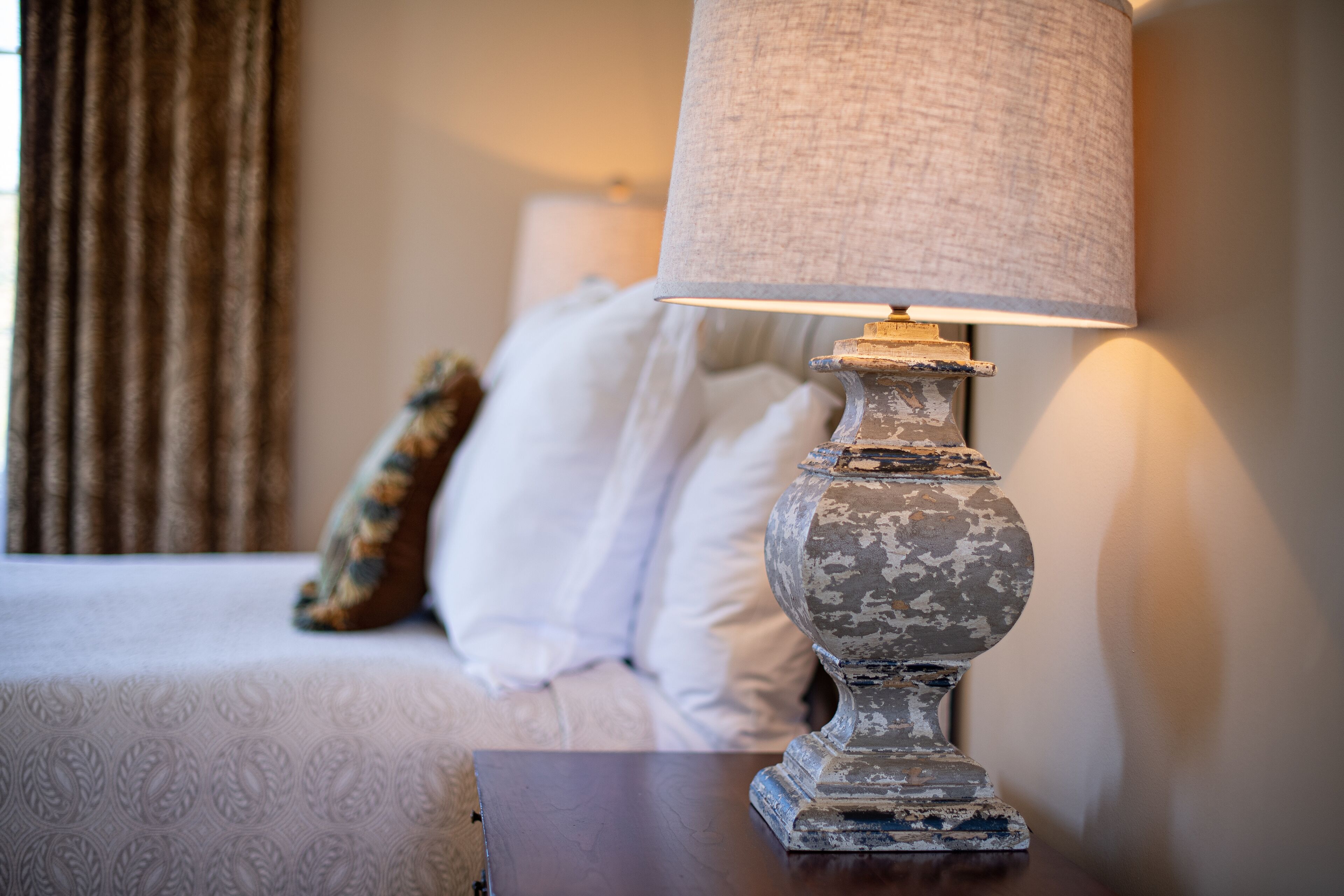 A bed with white linens and a decorative pillow, a wooden nightstand with a lamp, and a window with curtains.