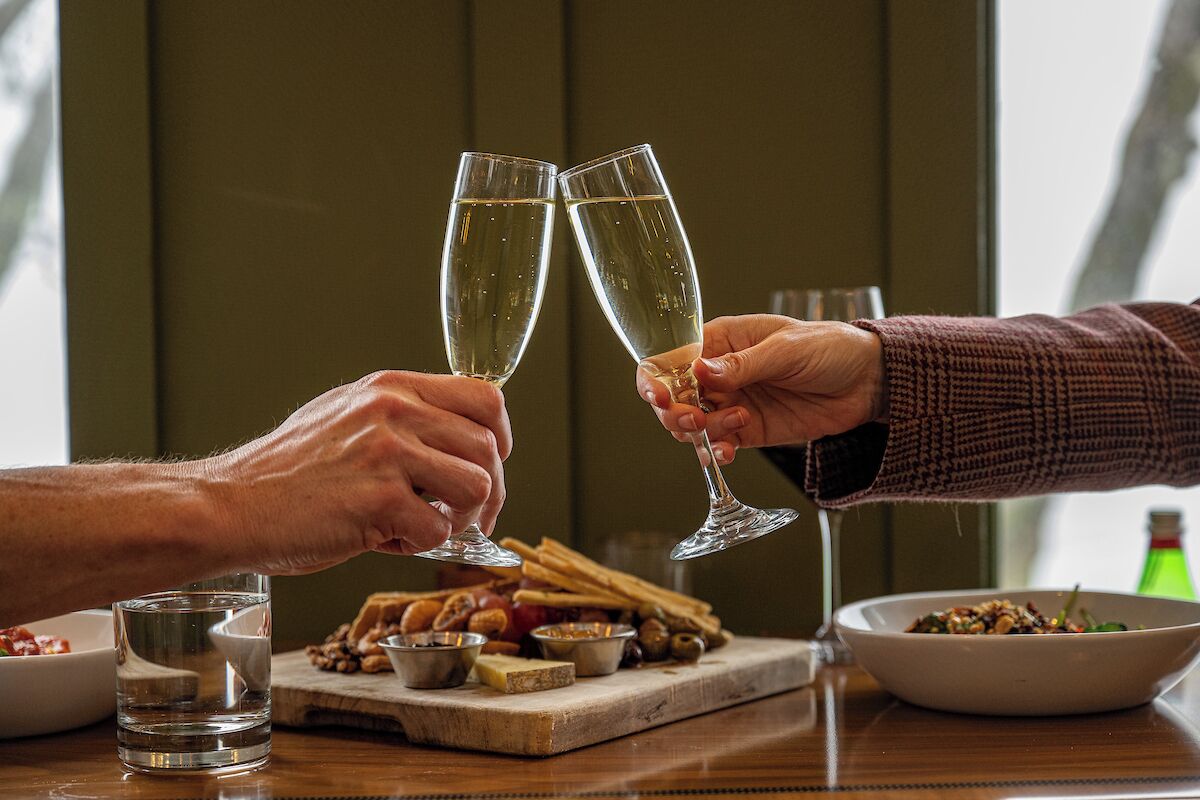 Zwei Hände stoßen Gläser Champagner an, eine Schale mit Nüssen und ein Glas Wasser stehen auf einem Holztisch.