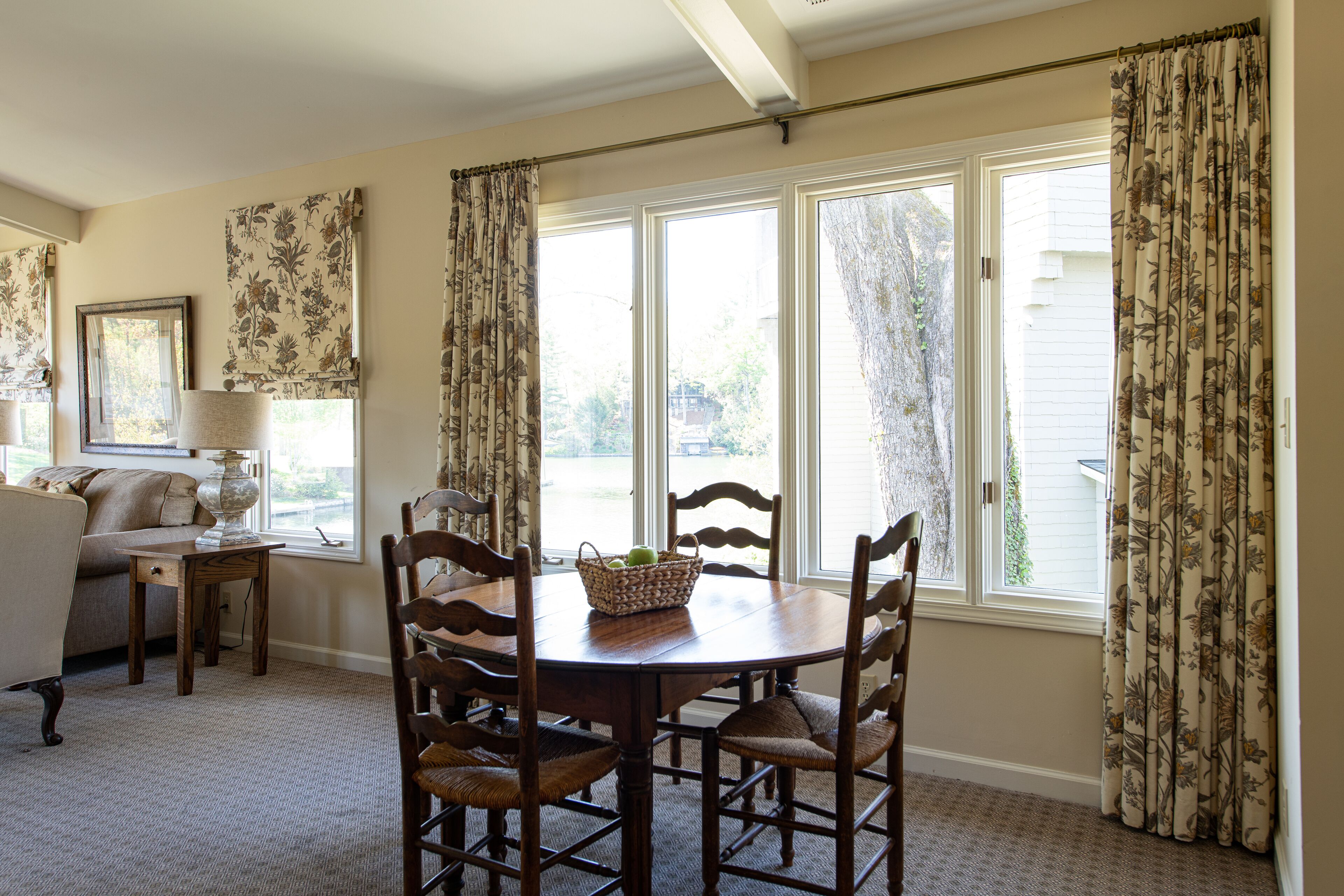 A dining area with a wooden table and chairs, a basket of fruit, and a window with curtains.