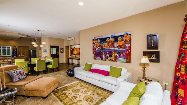 A spacious living room with a white sofa, green cushions, a colorful painting, and a dining area with green chairs.