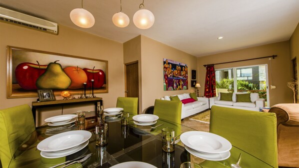 A dining area with a glass table set for four, surrounded by green chairs. There is a painting of fruits on the wall above the table.