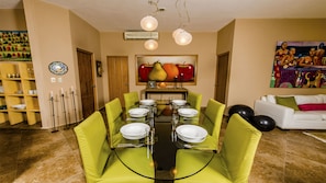 A dining area with a glass table surrounded by green chairs, a painting of fruits on the wall, and a white sofa in the background.