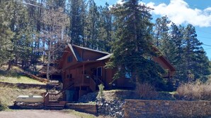 A log cabin with a porch and a stone wall in a forested area. - In town home- above the 1880 train.  Walk to Main St!— recently updated! (Hill City)