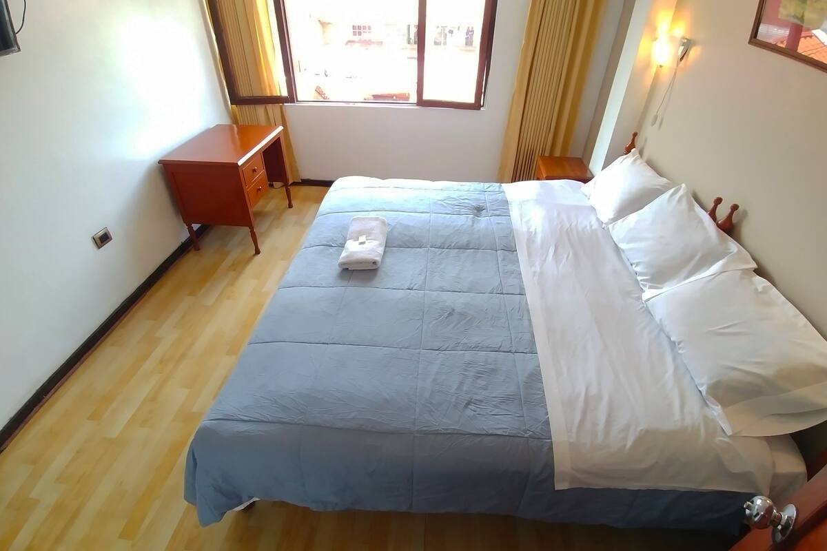 Ein ordentlich bezogenes Bett mit blauer und weißer Tagesdecke, ein Nachttisch aus Holz, ein kleiner Schreibtisch und ein Fenster mit Vorhängen.