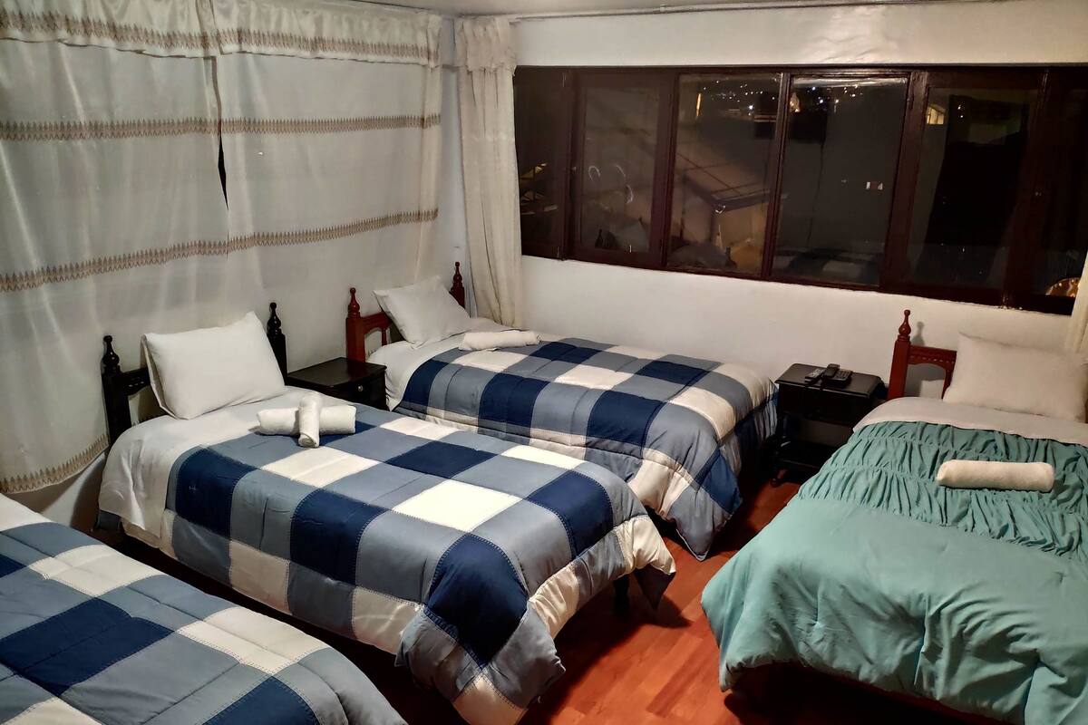 Ein Hotelzimmer mit drei Betten, jedes mit einer blau-weiß karierten Tagesdecke. Ein Fenster bietet Blick ins Freie, und der Boden ist mit Holz ausgelegt.