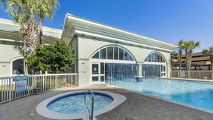 Une piscine avec un jacuzzi, entourée d’une clôture blanche, de palmiers et d’un bâtiment doté de fenêtres en arc de cercle.