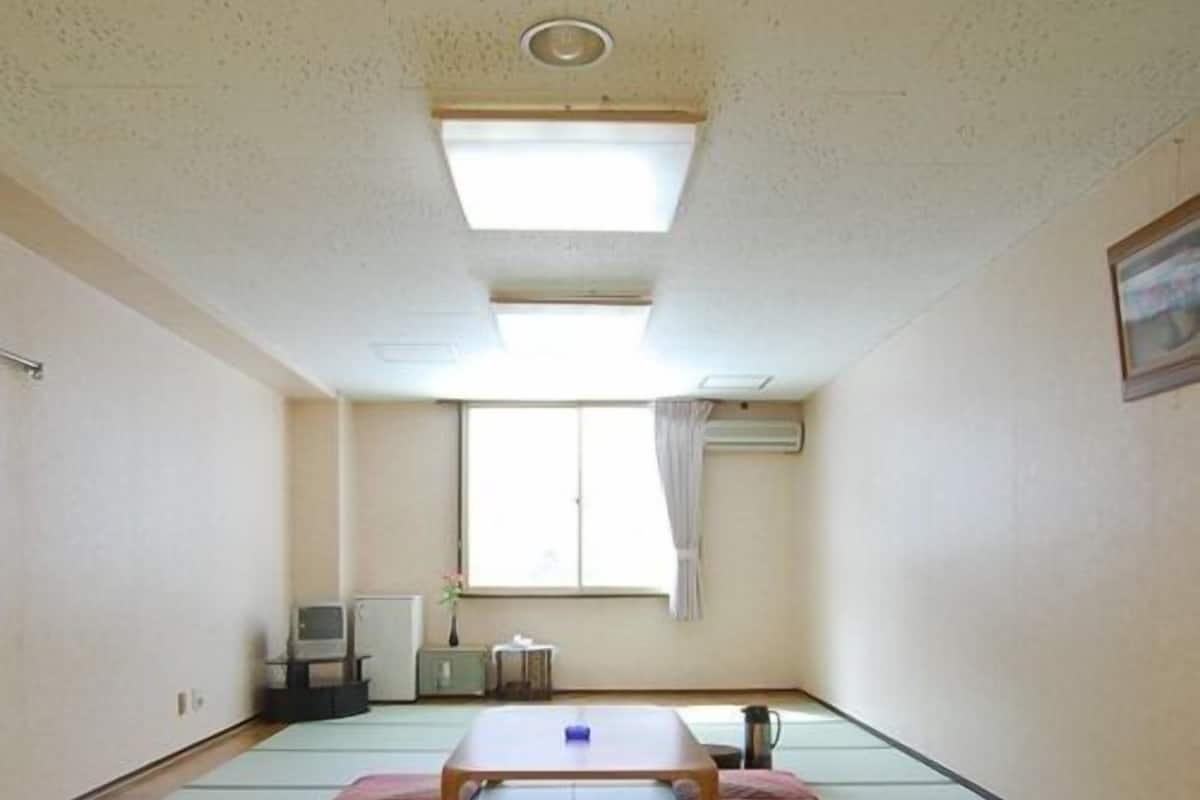 Una habitación con esteras de tatami, una mesa baja, un televisor, un radiador y una ventana con cortinas.