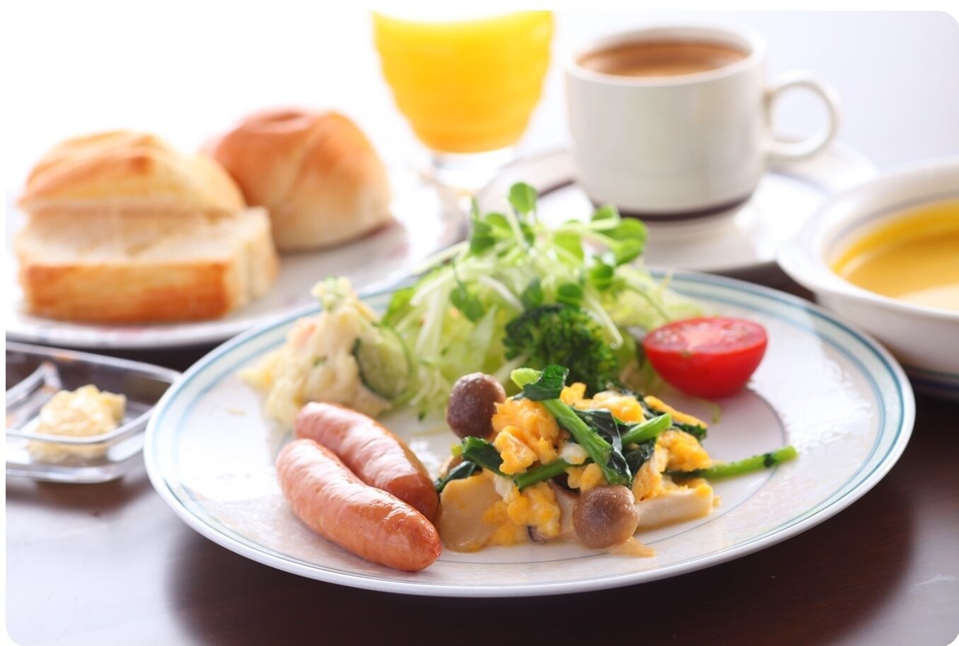 A breakfast plate with sausages, scrambled eggs, and greens, accompanied by a glass of orange juice, a cup of coffee, and a bowl of soup.