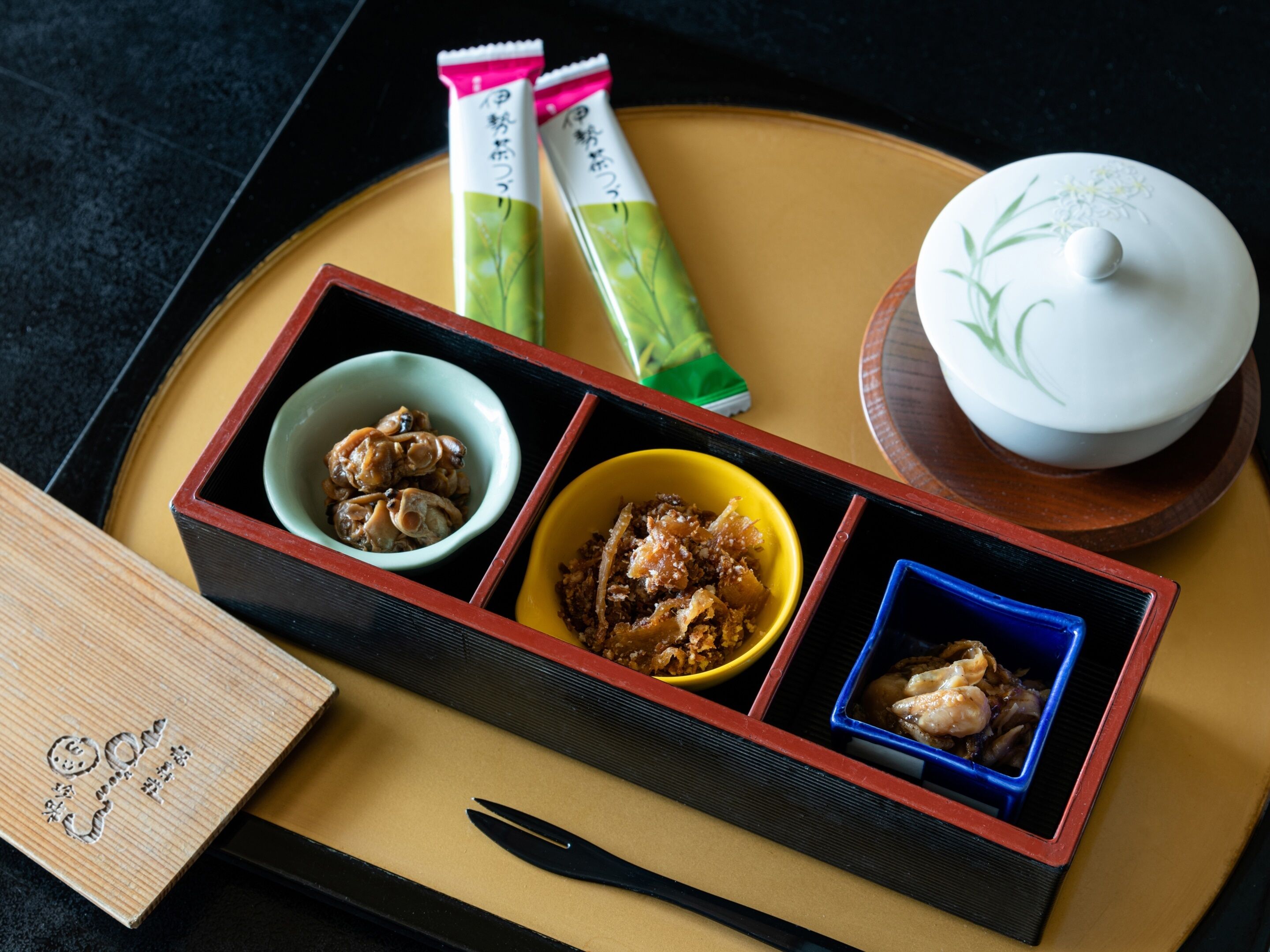 Un repas japonais traditionnel, composé de trois plats présentés dans un plateau en bois, accompagné de baguettes et d’un service à thé.