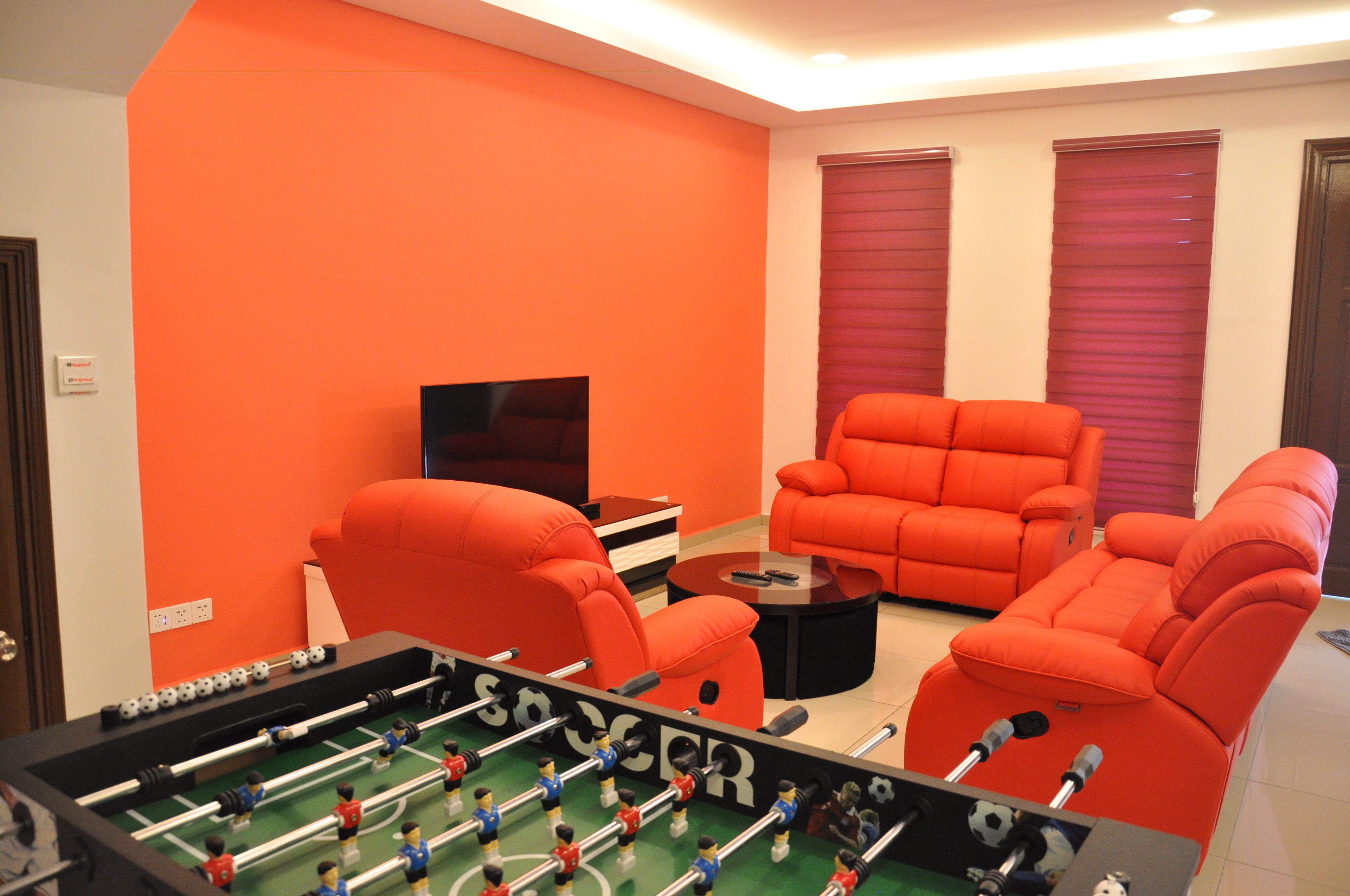 Een kamer met een tafelvoetbaltafel, twee oranje banken, een televisie en een raam met gesloten jaloezieën.