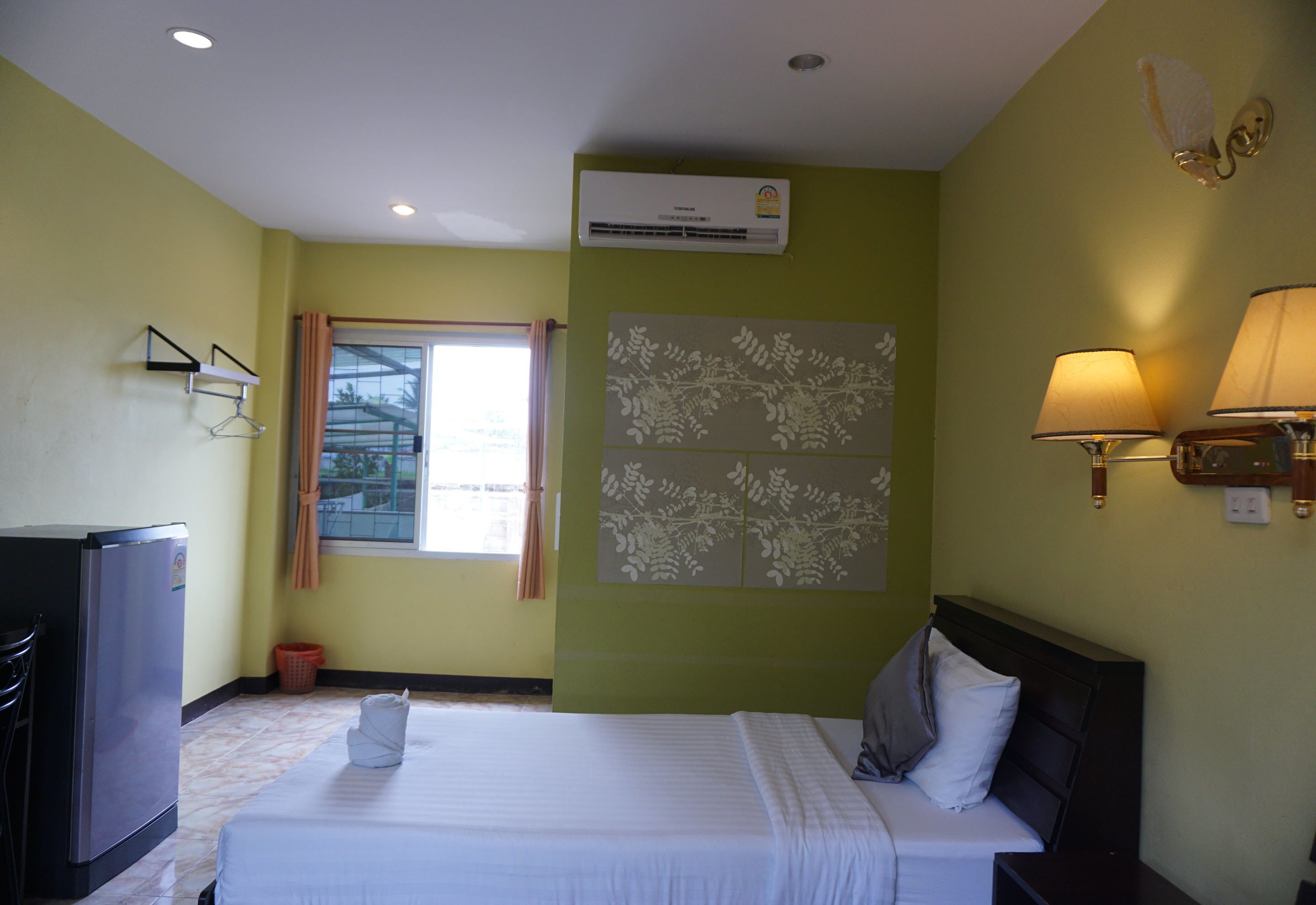 A hotel room with a bed, a desk, an air conditioner, a window with curtains, and wall-mounted lamps.