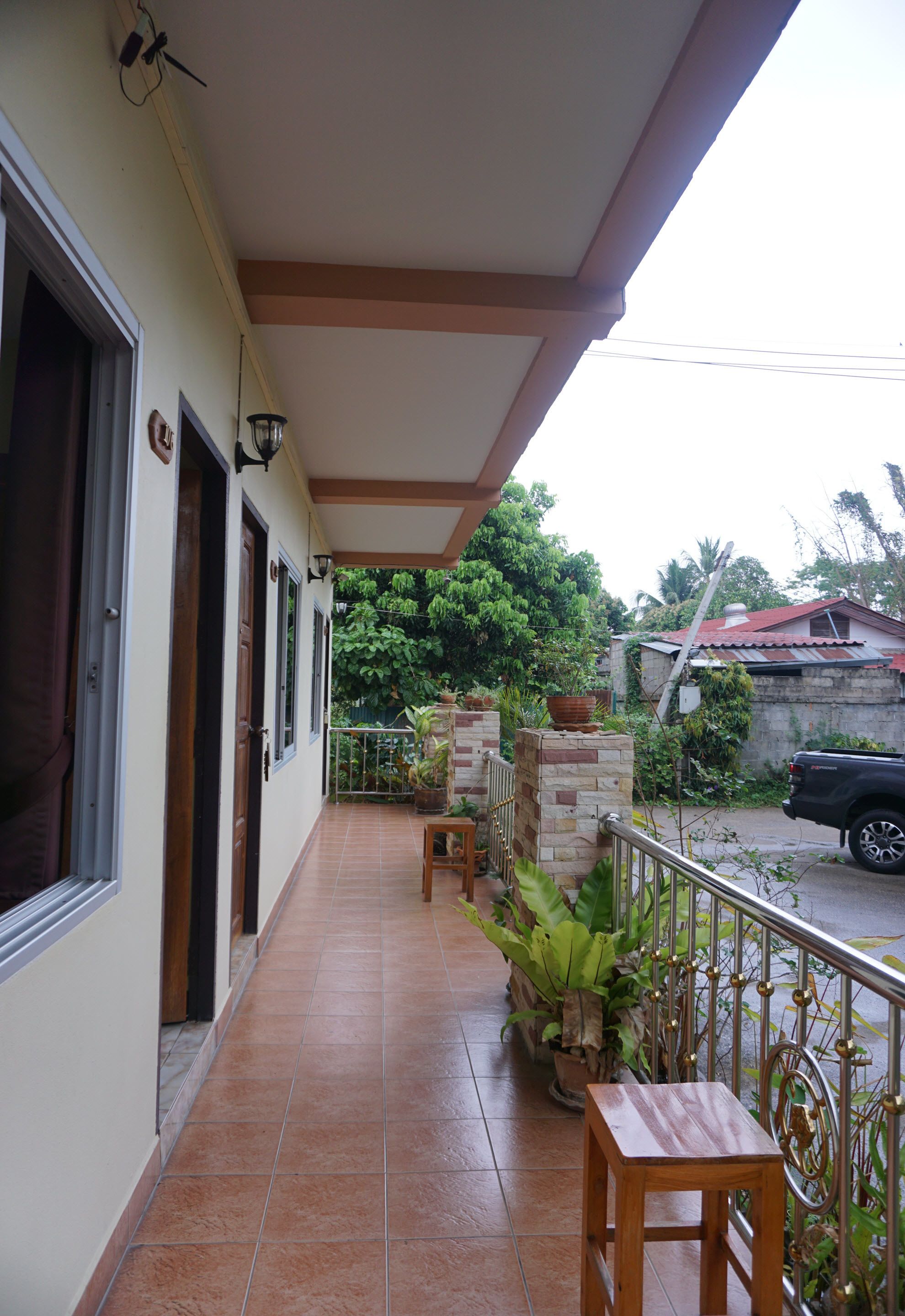 A covered balcony with tiled flooring, potted plants, and a metal railing.