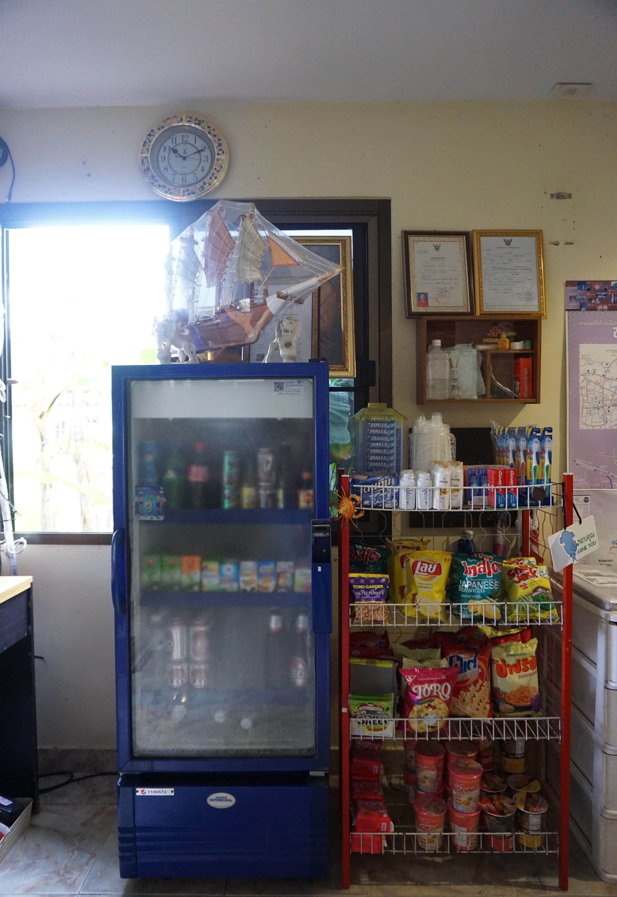 A small convenience store with a refrigerator, shelves stocked with snacks, and a clock on the wall.