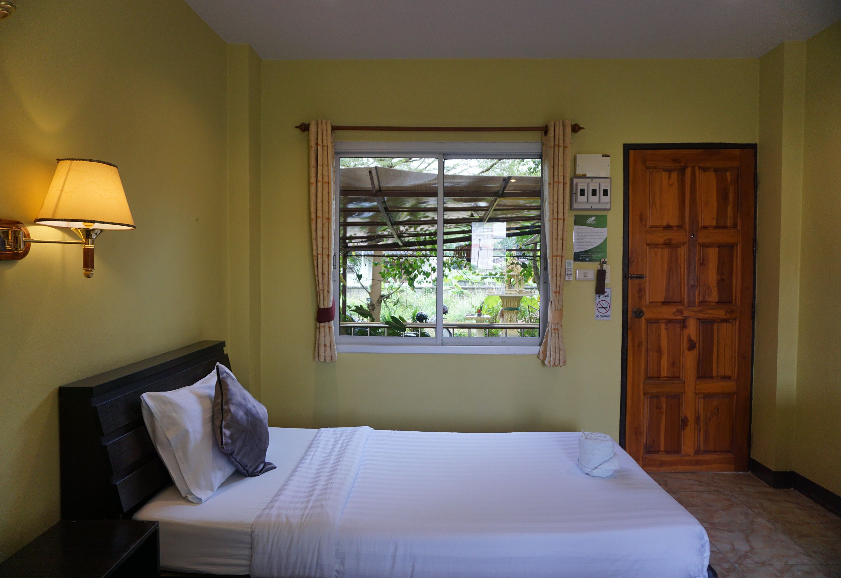 A hotel room with a bed, a nightstand, a lamp, a window with curtains, and a wooden door.
