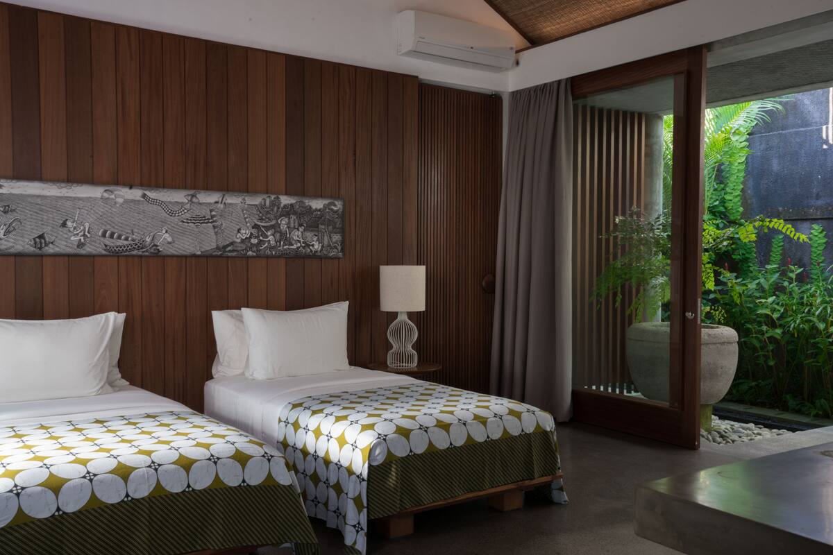 Une chambre d’hôtel avec deux lits, une grande fenêtre avec des rideaux, un climatiseur fixé au mur et un décor mural en panneaux de bois.