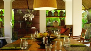A wooden dining table set with green placemats, clear glassware, and a bowl of fruit. - Atelier TE Designer Guesthouse (Denpasar)