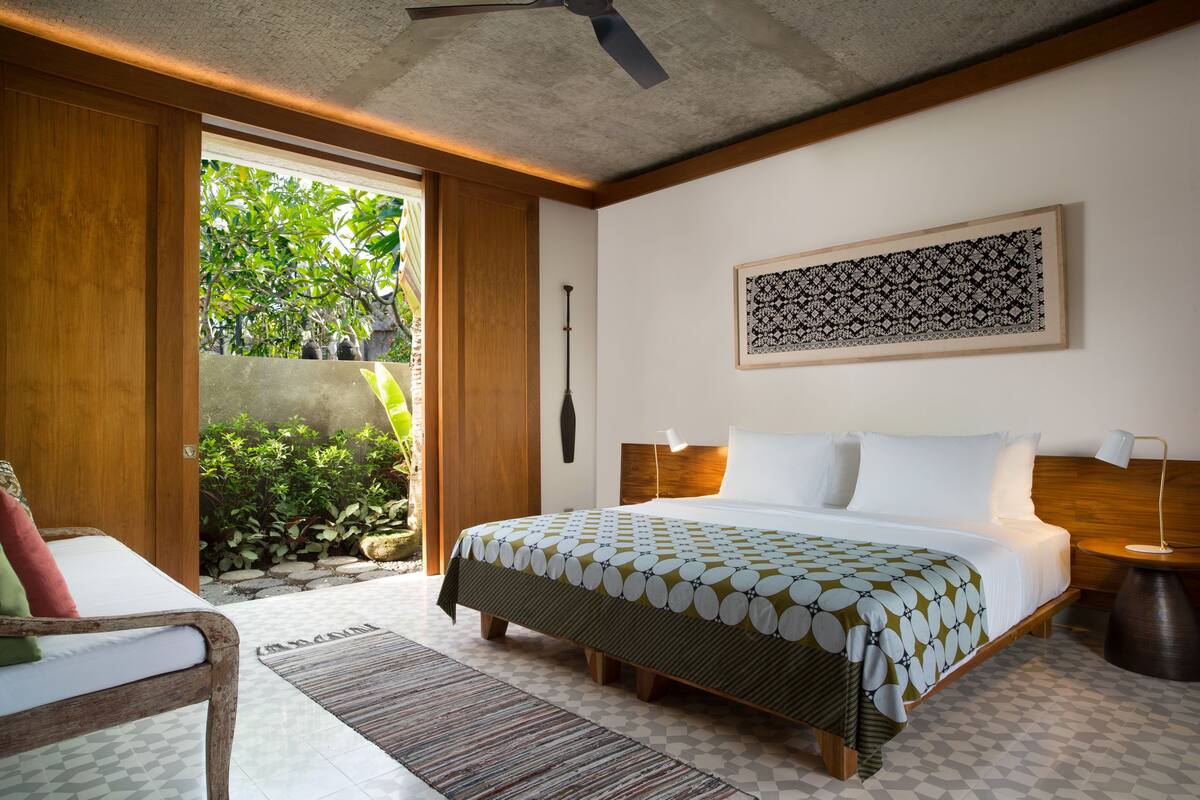 Une chambre à coucher avec un lit, une table de chevet, une lampe, un ventilateur de plafond et une vue sur la verdure à l’extérieur.