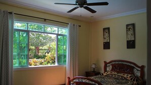 Un dormitorio con cama, ventilador de techo, ventana con cortinas y dos cuadros colgados en la pared.