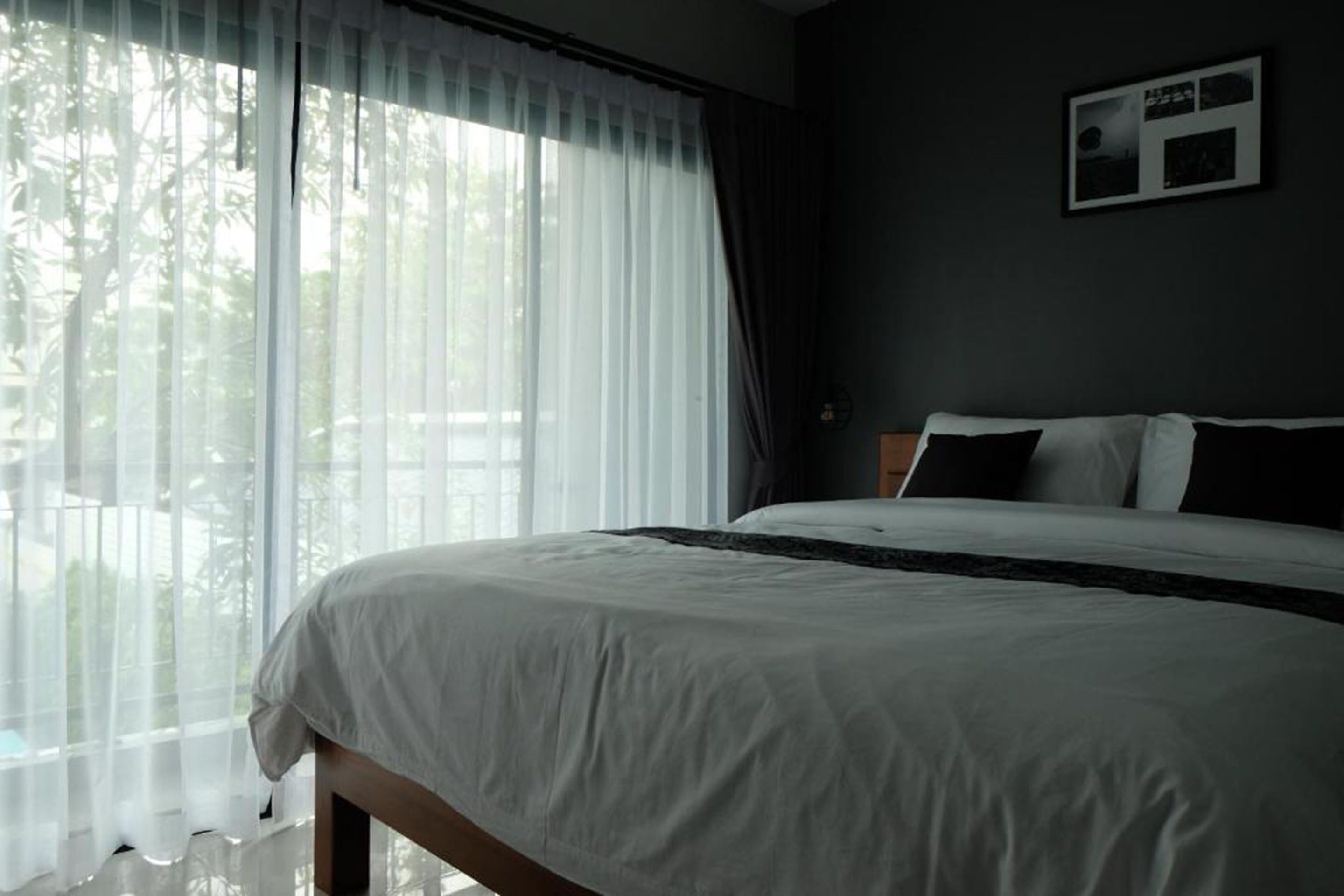 A bedroom with a large bed, white bedding, a view of trees through sheer curtains, and a framed picture on the wall.