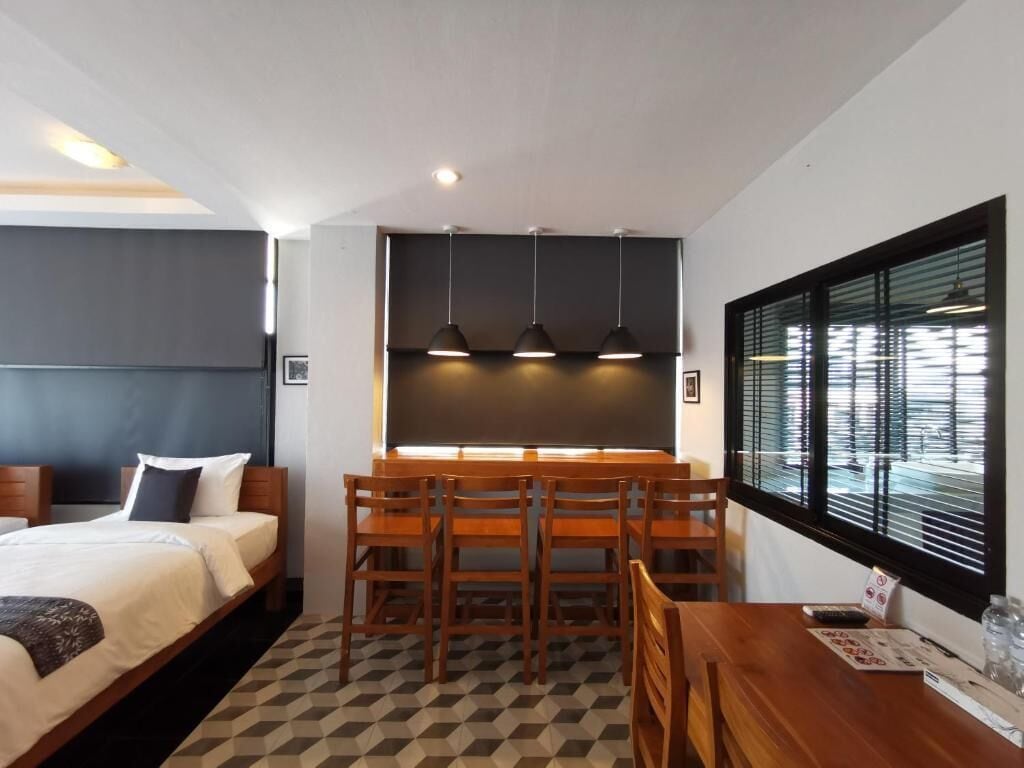 A hotel room with a bed, a desk, chairs, a blackboard, pendant lights, and a window with blinds.