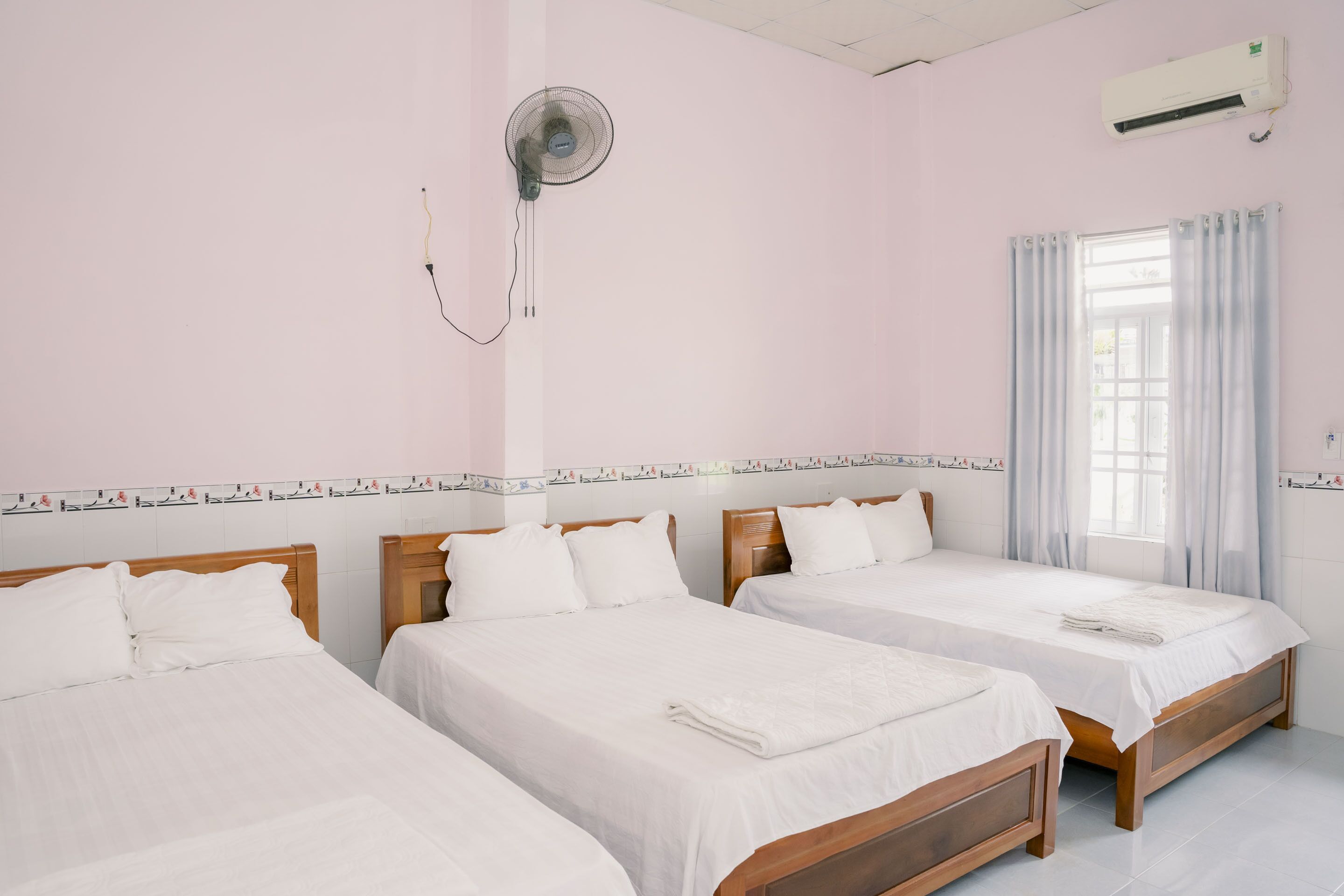 A hotel room with three single beds, a ceiling fan, an air conditioning unit, and a window with curtains.