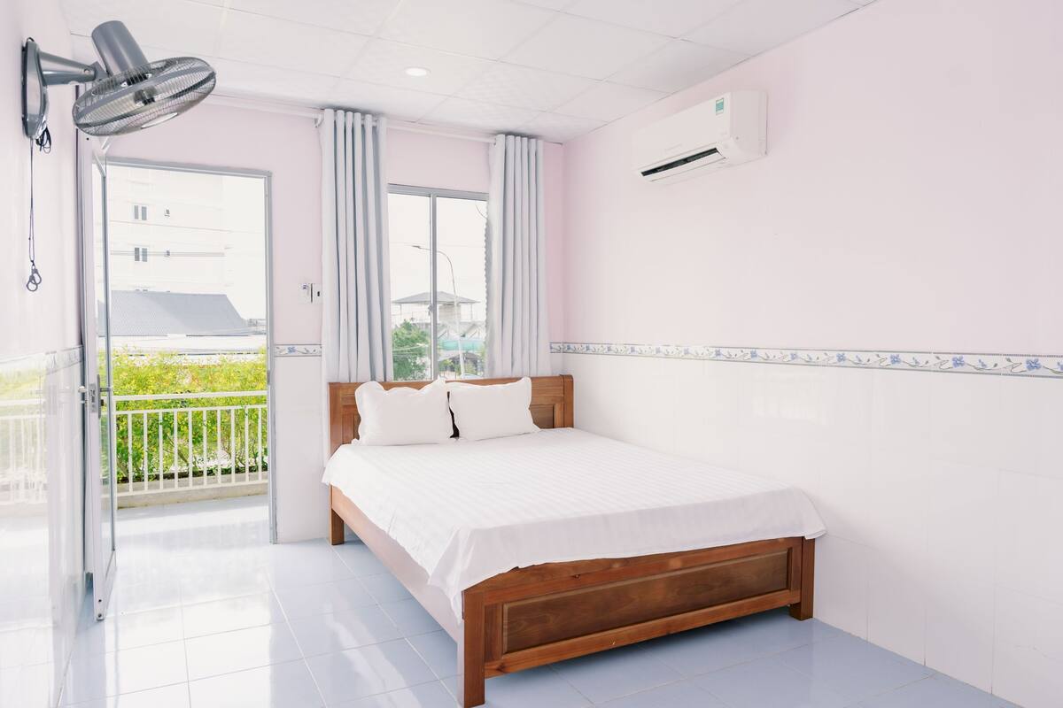 Une chambre avec un lit en bois, du linge de lit blanc, un ventilateur de plafond, un climatiseur et une vue sur la verdure, visible à travers la porte d’un balcon.