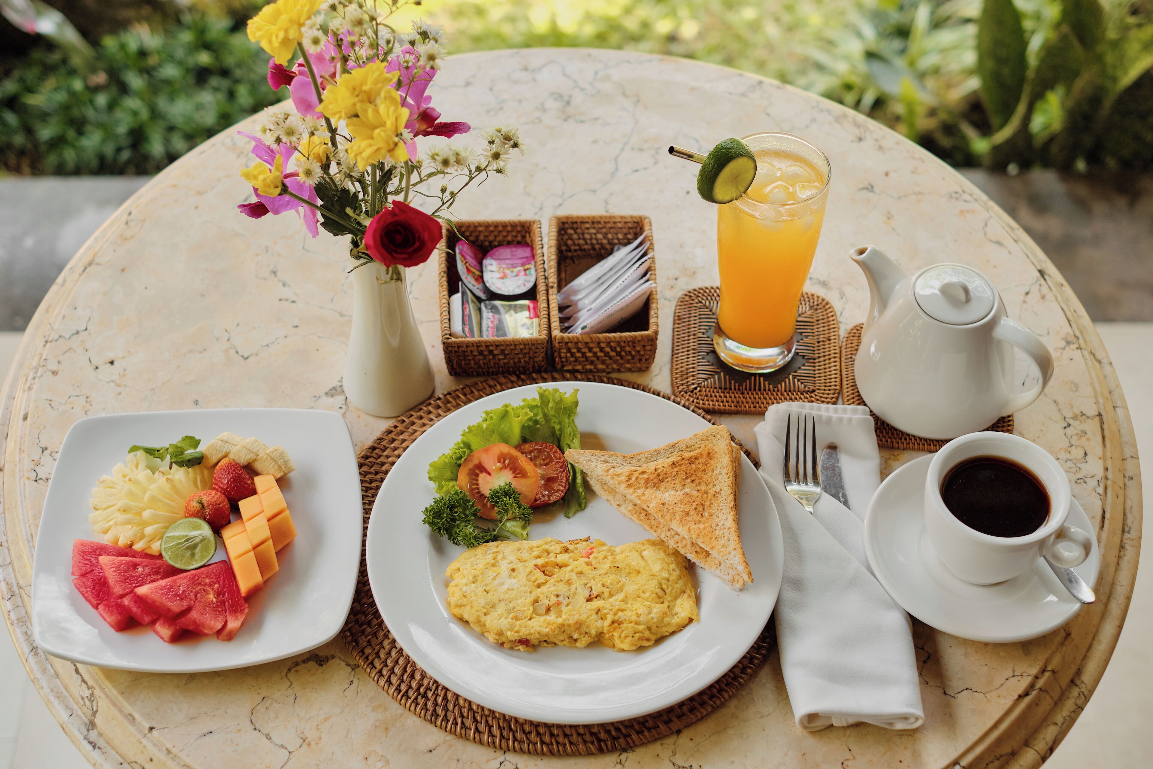 A breakfast setting with a plate of eggs, toast, and fruit, a glass of orange juice, a vase of flowers, and a teapot on a round table.