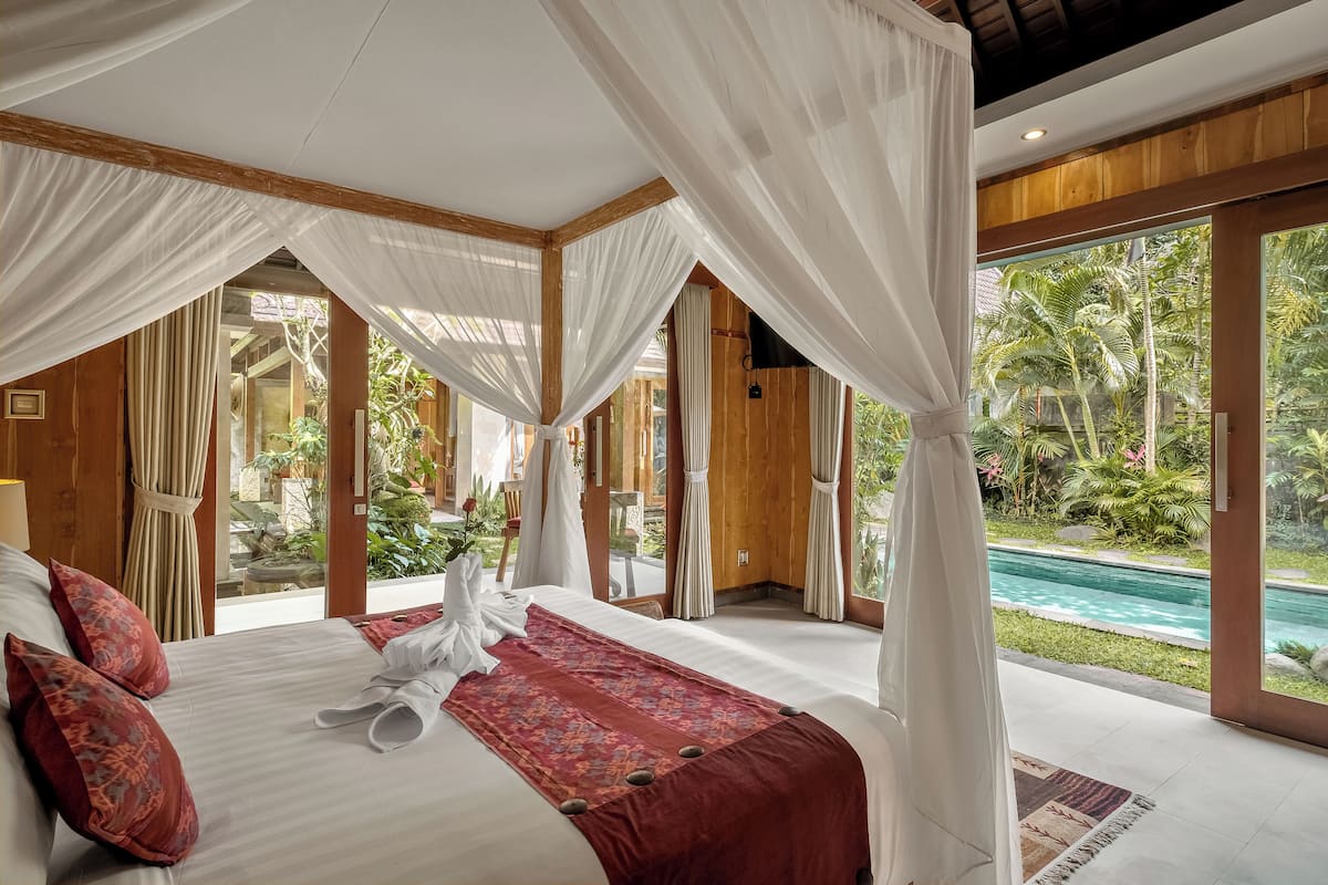 A spacious bedroom with a large bed, white curtains, and a view of a swimming pool.