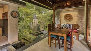 Una mesa de comedor de madera con sillas, una estatua de piedra y un exuberante jardín verde visible a través de una pared de vidrio.