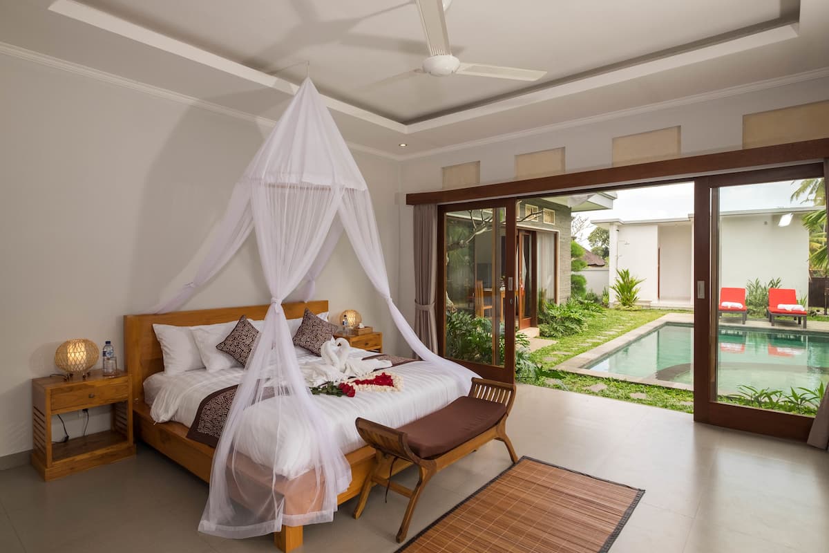 A bedroom with a canopy bed, a wooden nightstand, a bench, and a view of a swimming pool and garden.