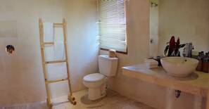 A bathroom with a toilet, a sink, a mirror, and a window with blinds. - Warisan Ubud Villa (Tegallalang)