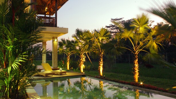 A pool area with lounge chairs, palm trees, and a building with a thatched roof. - Warisan Ubud Villa (Tegallalang)