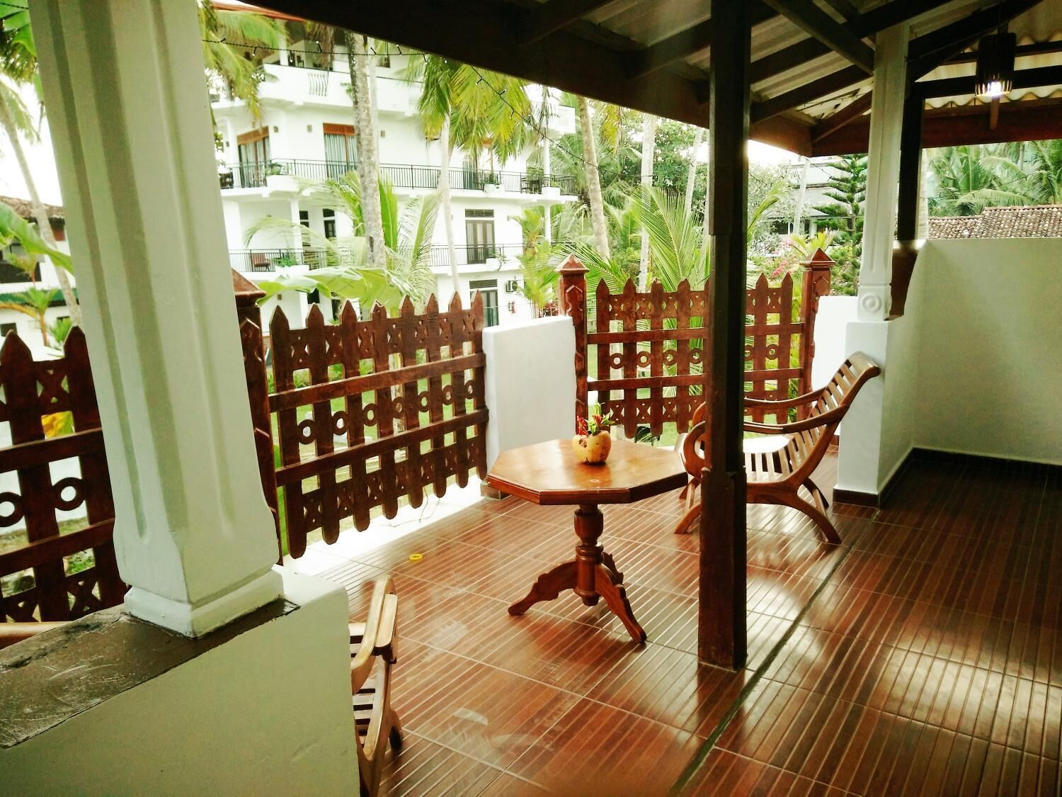 A covered outdoor area with a wooden table and chairs, a decorative fence, and a view of a white building with balconies and greenery.