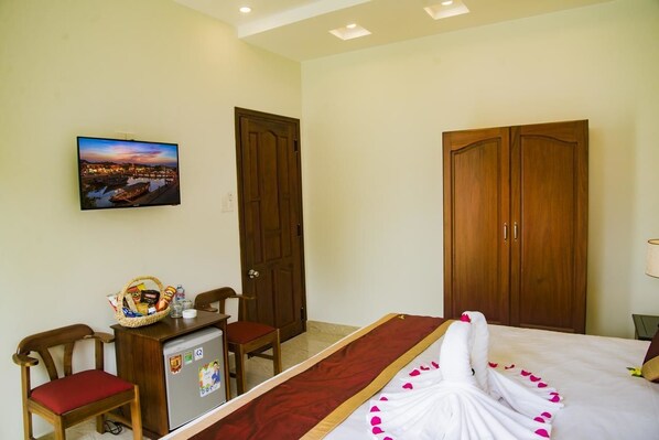 A hotel room with a bed, a desk with a refrigerator, a chair, a wooden wardrobe, and a wall-mounted TV displaying a cityscape. - Mi Kha Homestay (Da Nang)