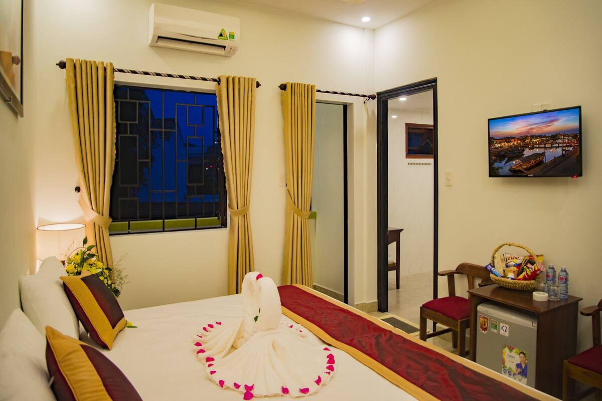 A hotel room with a bed, a TV, a desk with a basket, and a window with curtains.