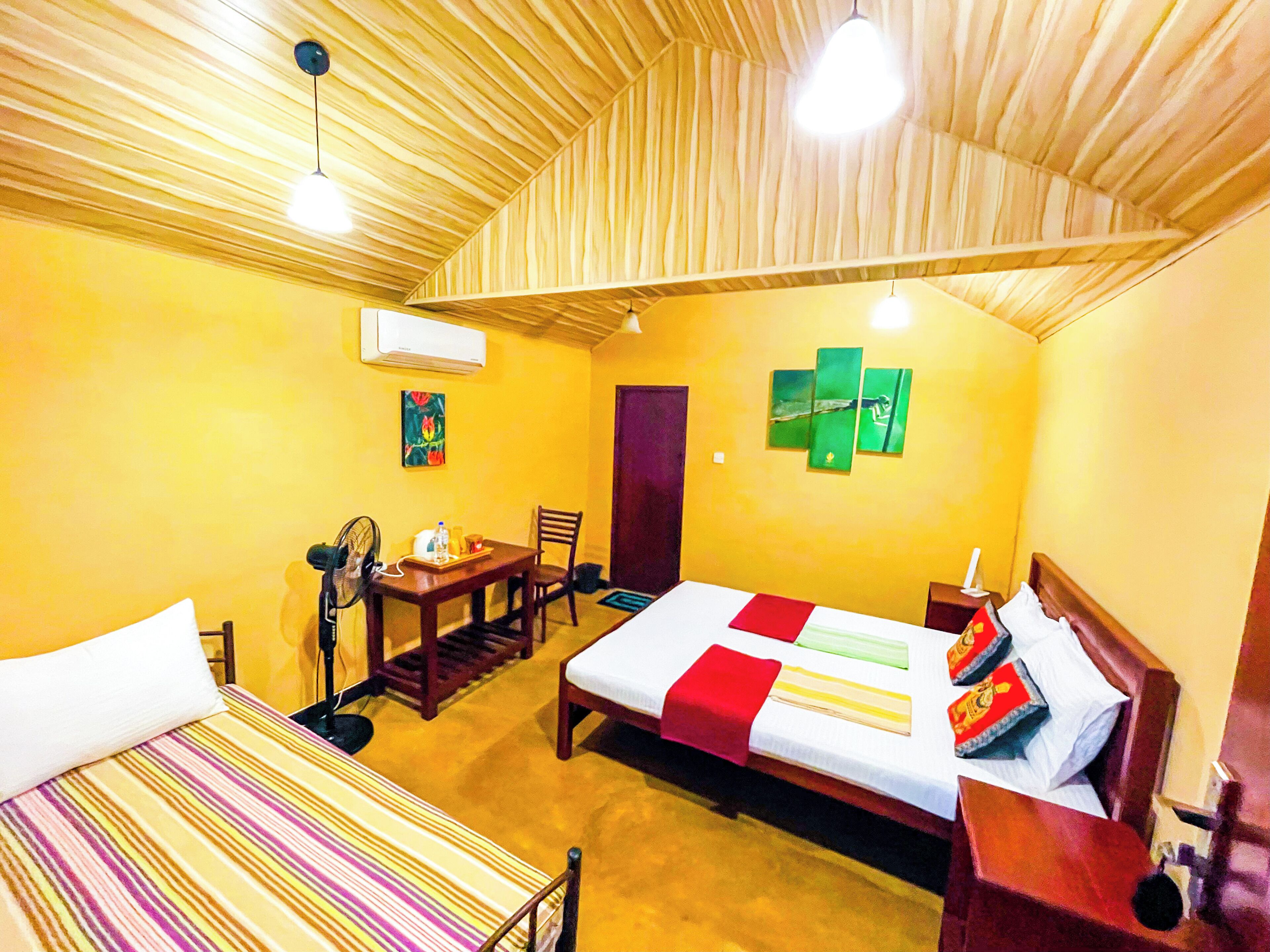 A room with a bed, a desk, a chair, and a fan. There is a door and a wall with a painting.