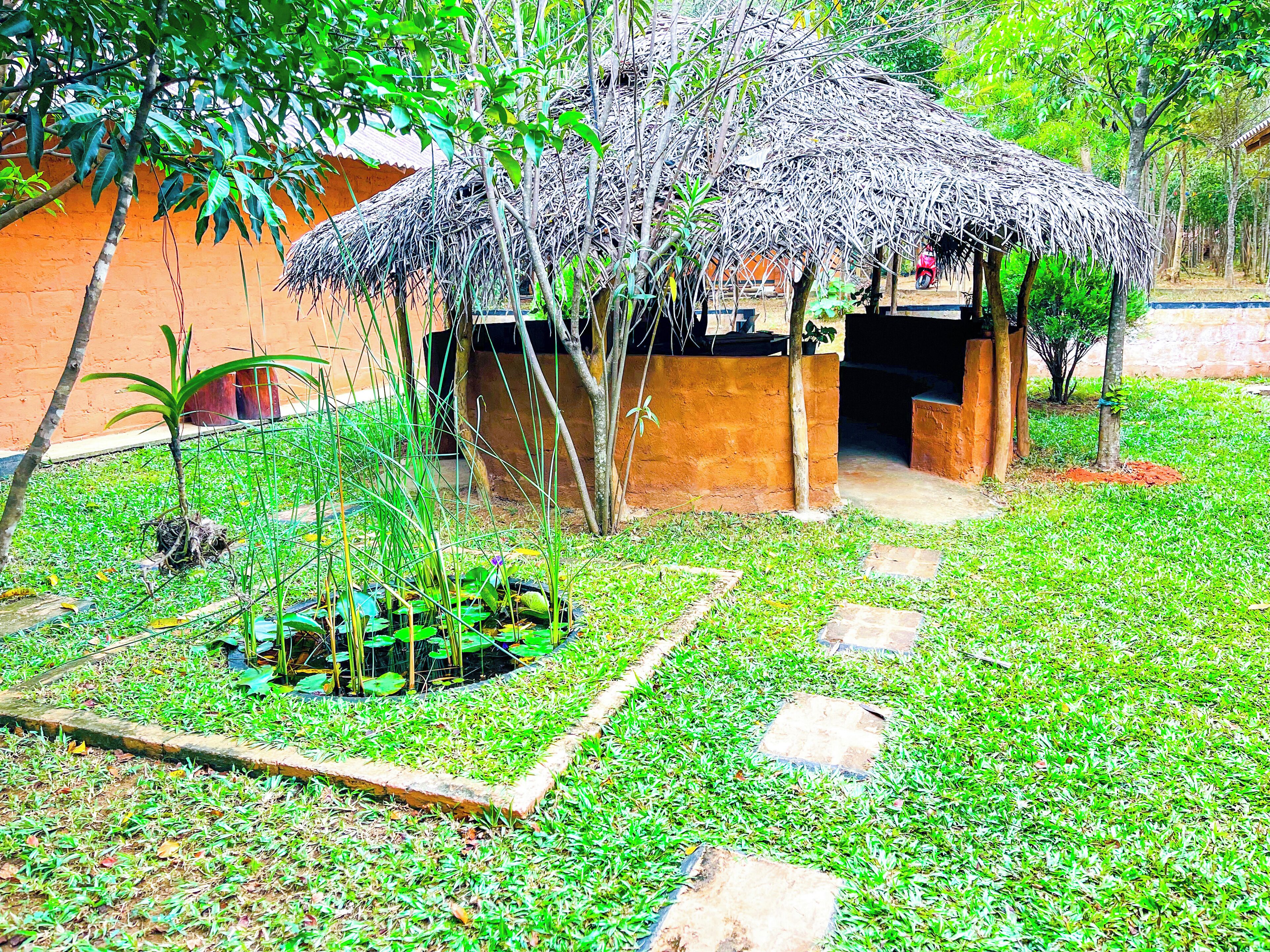 A thatched-roof structure with a garden area featuring a small pond, stepping stones, and various plants.