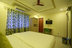 Una habitación de hotel con cama, ventilador de techo, unidad de aire acondicionado, televisión, una silla verde y un cesto de basura.
