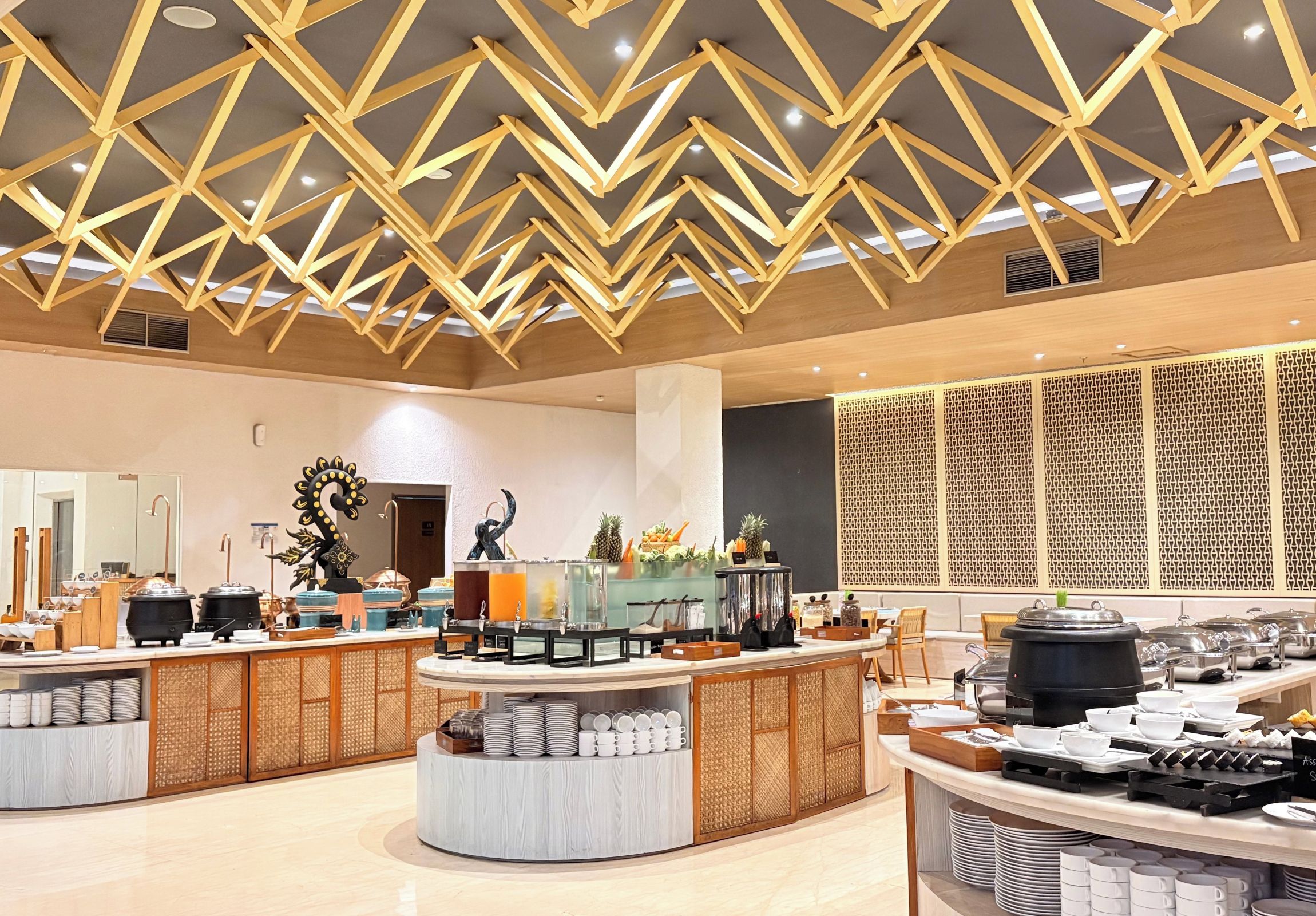 A modern hotel lobby with a central buffet area, decorative wall panels, and a geometric ceiling design.