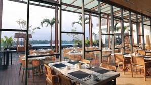 Un restaurant doté de grandes fenêtres offrant une vue sur des palmiers et un plan d’eau. Les tables sont dressées avec des nappes blanches et des chaises en osier.