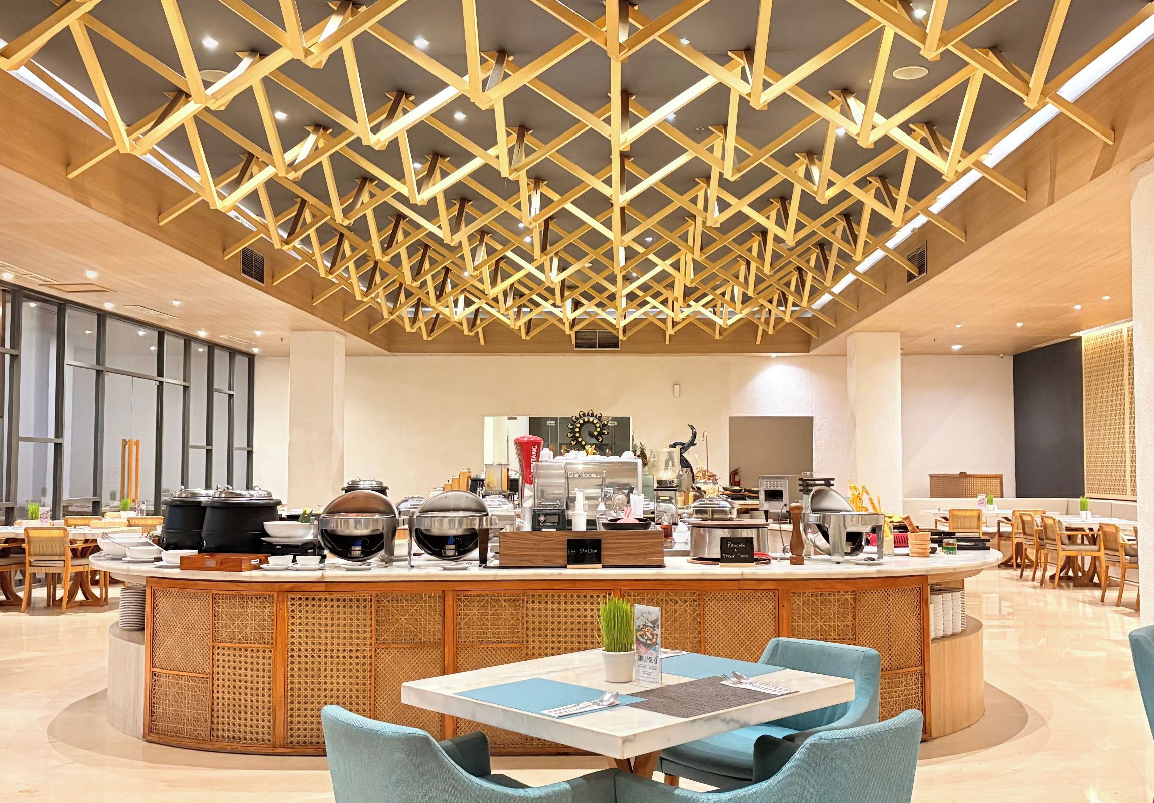 Un comedor moderno con barra central, mesas redondas y un techo alto con diseño geométrico de madera.