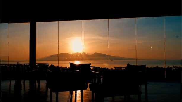 Une vue sur le coucher de soleil depuis une chambre dotée de grandes fenêtres, avec une table et des chaises disposées face à la vue.