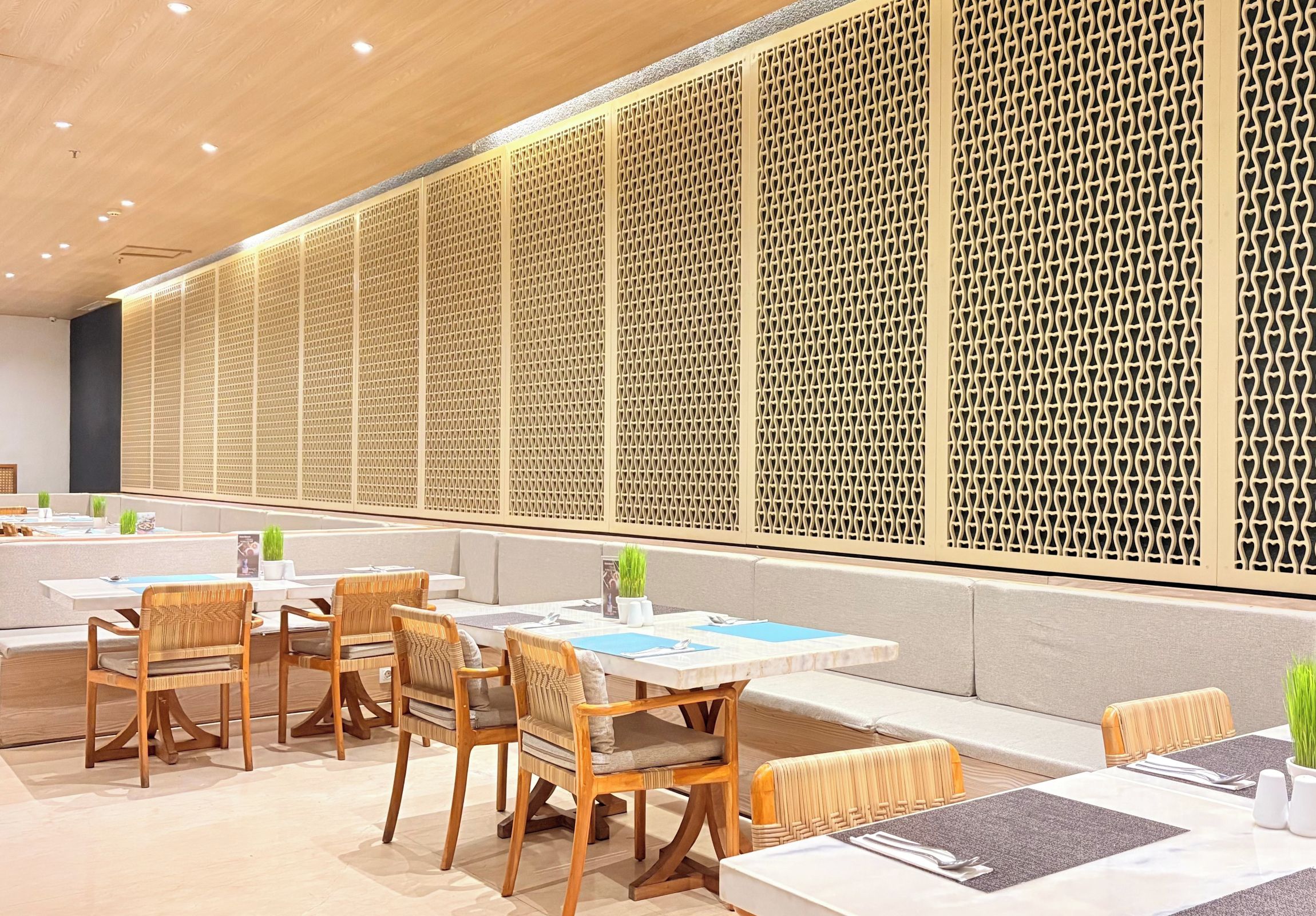 Un área de comedor moderna con mesas y sillas de madera, un muro con diseño y un techo con iluminación empotrada.
