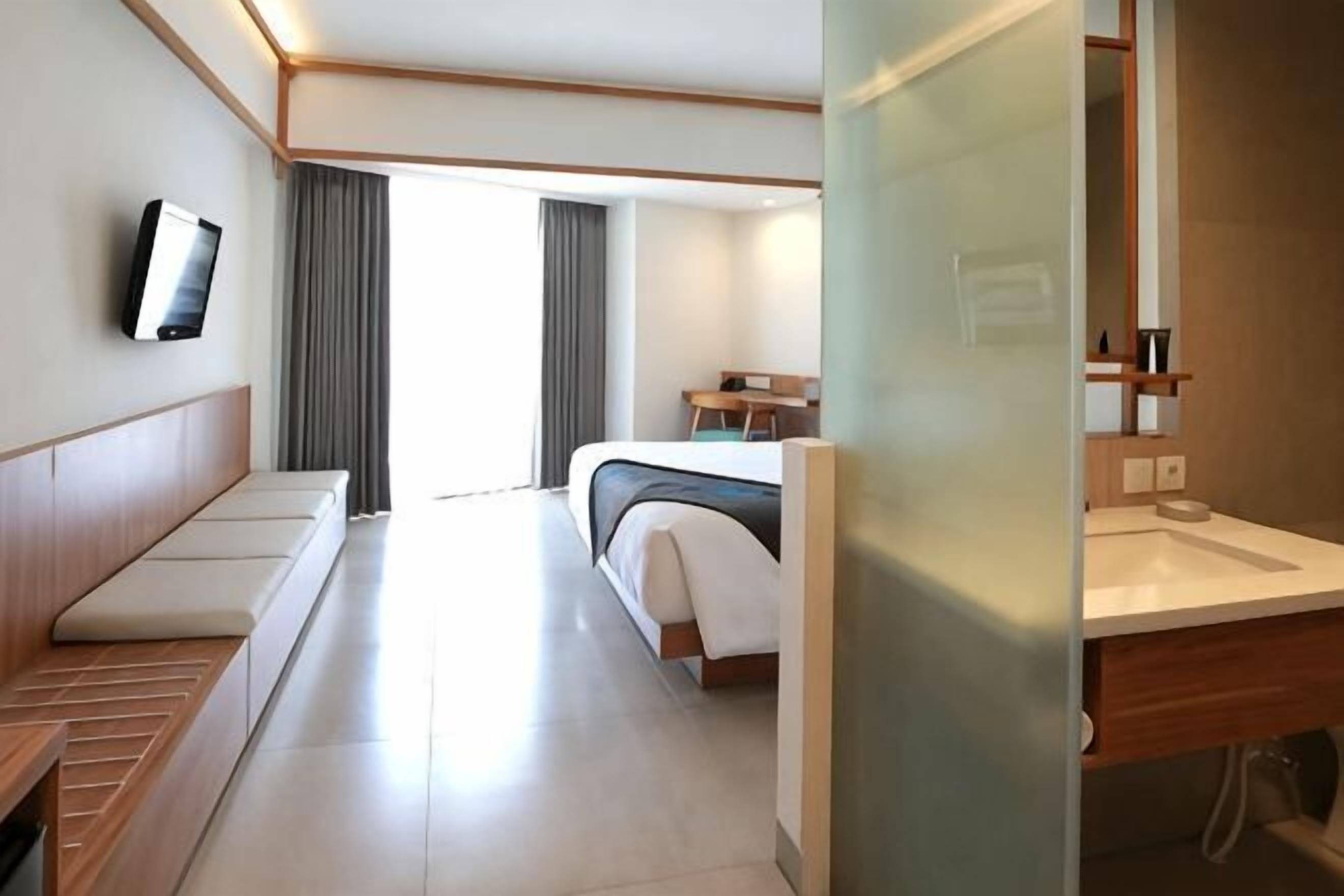 A hotel room with a bed, a seating area, a television, a bathroom with a sink, and a window with curtains.